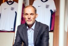 A man in a suit seated before a wall adorned with various England football shirts.