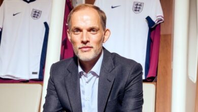 A man in a suit seated before a wall adorned with various England football shirts.