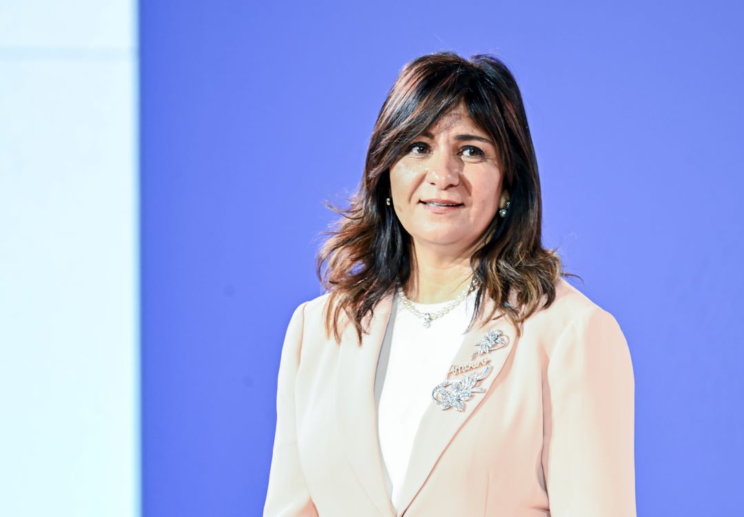 "A person in a light-colored blazer with a brooch stands against a blue background."