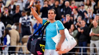 “Tennis player with obscured face waving to the crowd while carrying multiple bags, indicating a possible victory or farewell, with spectators’ faces pixelated for privacy.”