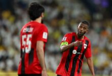 Two men wearing red and black soccer uniforms stand together on a field