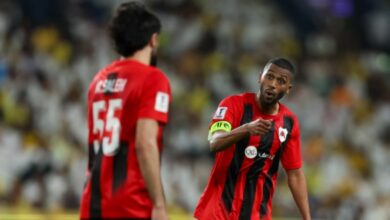 Two men wearing red and black soccer uniforms stand together on a field